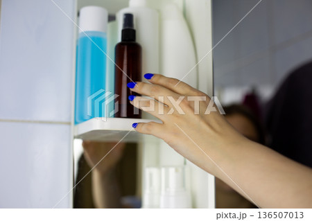 Hand with brightly manicured hand reaches for skincare bottle on bathroom shelf. Concept of skin and body care, health through cleaning products. Special bathroom products Hand with brightly manicured hand reaches for skincare bottle on bathroom shelf. Concept of skin and body care, health through cleaning products. Special bathroom products 136507013