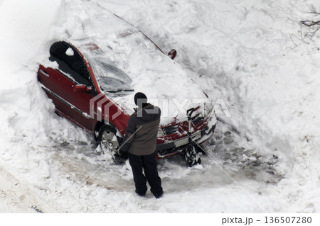 Adult male clearing deep accumulation of white snow from top and sides of red passenger car using shovel after heavy winter snowfall Adult male clearing deep accumulation of white snow from top and sides of red passenger car using shovel after heavy winter snowfall 136507280