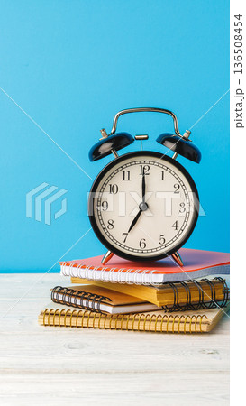 Clock sits on notebooks against a blue backdrop during daytime in a study area Clock sits on notebooks against a blue backdrop during daytime in a study area 136508454