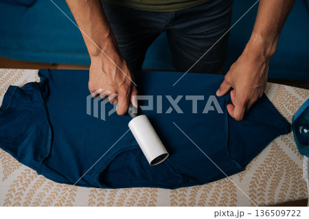 Closeup top view of unrecognizable man using sticky lint roller to remove dust and lint from blue t-shirt on ironing board, with steam iron nearby, preparing to iron and care for clothes at home. Closeup top view of unrecognizable man using sticky lint roller to remove dust and lint from blue t-shirt on ironing board, with steam iron nearby, preparing to iron and care for clothes at home. 136509722