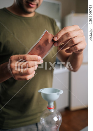 Vertical cropped shot of gardener pouring fertilizer from sachet into bottle using funnel, preparing nutrient solution for plants, demonstrating simple yet effective method for plant care at home. Vertical cropped shot of gardener pouring fertilizer from sachet into bottle using funnel, preparing nutrient solution for plants, demonstrating simple yet effective method for plant care at home. 136509748