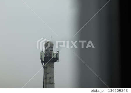 Industrial Chimney with Communication Antennas. Industrial concrete chimney with smoke and telecommunication antennas under a grey sky, urban pollution concept Industrial Chimney with Communication Antennas. Industrial concrete chimney with smoke and telecommunication antennas under a grey sky, urban pollution concept 136509848