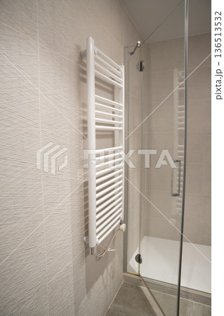 Modern bathroom featuring a white heated towel rail radiator mounted on textured ceramic wall tiles, providing comfort and contemporary design beside a glass shower door Modern bathroom featuring a white heated towel rail radiator mounted on textured ceramic wall tiles, providing comfort and contemporary design beside a glass shower door 136513532