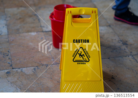 Bright yellow wet floor warning sign on tiled floor with red bucket in background, highlighting cleaning in progress and safety precautions to prevent slips, falls, and accidents 136513554