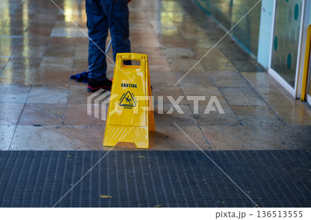 Yellow caution sign warning about a wet floor in a public building, a worker mopping the shiny tiled surface nearby, emphasizing cleaning, safety, and hygiene 136513555