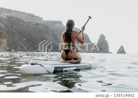 Woman Paddleboarding Ocean Coastline - A woman kneels on a paddleboard in the ocean, facing a rocky coastline. The sun is shining and the water is calm. Woman Paddleboarding Ocean Coastline - A woman kneels on a paddleboard in the ocean, facing a rocky coastline. The sun is shining and the water is calm. 136514982