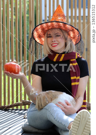 A beautiful girl is sitting on the terrace with a dog, holding a pumpkin in her hand. 136515312