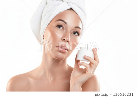 Beautiful Caucasian woman with white towel wrapped around her head. She holds cream jar in hand, isolated on white. 136516231