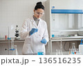 Female scientist in white coat and safety goggles conducting a chemical experiment in a laboratory. 136516235