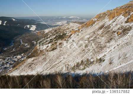 Torgashinsky ridge, Krasnoyarsk, Siberia. Winter landscape of a snow covered mountain slope with dry trees below and a small village in the valley. Nature photography Torgashinsky ridge, Krasnoyarsk, Siberia. Winter landscape of a snow covered mountain slope with dry trees below and a small village in the valley. Nature photography 136517882