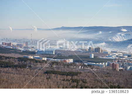 Panoramic aerial view of Krasnoyarsk city in winter. Urban landscape with buildings, forest, and mountains in fog. Environmental concept. 136518623