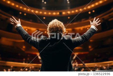 Back view of conductor with raised arms in concert hall, dramatic stage lighting and theater seating creating atmospheric performance scene Back view of conductor with raised arms in concert hall, dramatic stage lighting and theater seating creating atmospheric performance scene 136522936