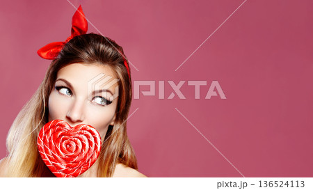 Woman Pin-Up Enjoying Heart Lollipop, Playful Portrait With Red Bandana, Glossy Lipstick, Pink Background, CloseUp Framing, Flirtatious Sideways Gaze, Vintage Style, Commercial Beauty, Valentines Day 136524113
