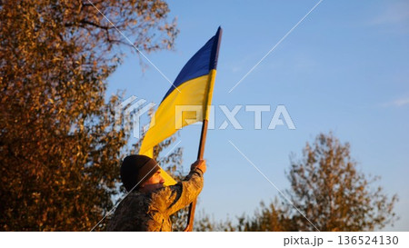Male military in uniform waving flag of Ukraine at countryside. Young soldier of ukrainian army lifting blue-yellow banner as symbol of victory against russian aggression. Invasion resistance concept Male military in uniform waving flag of Ukraine at countryside. Young soldier of ukrainian army lifting blue-yellow banner as symbol of victory against russian aggression. Invasion resistance concept 136524130