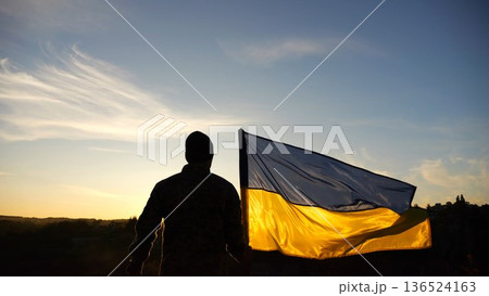 Silhouette of ukrainian army man stands with national banner at countryside on sunset. Male soldier in military uniform with Ukraine flag as symbol of victory against russian aggression. Slow motion Silhouette of ukrainian army man stands with national banner at countryside on sunset. Male soldier in military uniform with Ukraine flag as symbol of victory against russian aggression. Slow motion 136524163
