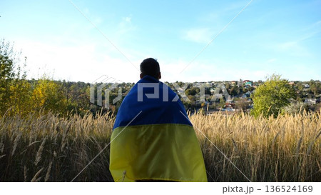 Military man in uniform stands with flag of Ukraine on shoulders at countryside. Portrait of male ukrainian soldier with national banner in honor of victory against russian aggression. End of war Military man in uniform stands with flag of Ukraine on shoulders at countryside. Portrait of male ukrainian soldier with national banner in honor of victory against russian aggression. End of war 136524169