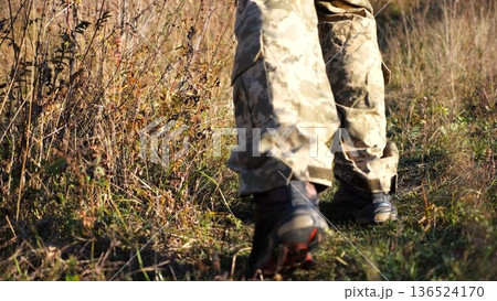 Low view to male feet of ukrainian soldiers going along trail at countryside. Legs of young army man walking through dry grass at sunny day. War in Ukraine. Slow motion Low view to male feet of ukrainian soldiers going along trail at countryside. Legs of young army man walking through dry grass at sunny day. War in Ukraine. Slow motion 136524170