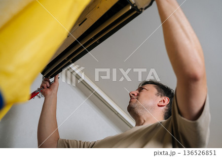 Low-angle view of technician carefully disassembles and repairs wall-mounted air conditioning unit, using screwdriver to adjust internal components and ensure proper functionality. Low-angle view of technician carefully disassembles and repairs wall-mounted air conditioning unit, using screwdriver to adjust internal components and ensure proper functionality. 136526851
