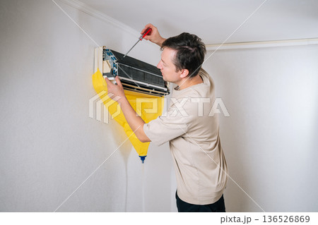 Back view of HVAC technician unscrewing air conditioning unit during maintenance or repair intervention, using screwdriver and yellow protective sheet. Concept of air condition maintenance service. Back view of HVAC technician unscrewing air conditioning unit during maintenance or repair intervention, using screwdriver and yellow protective sheet. Concept of air condition maintenance service. 136526869