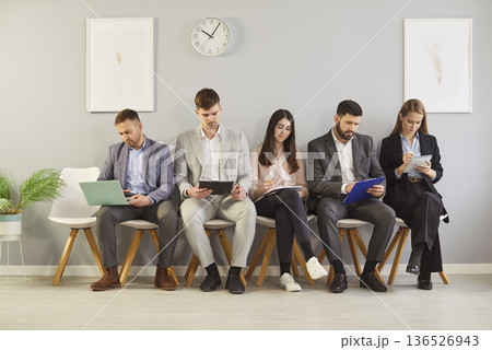Group of diverse professionals sitting in waiting area, using devices and preparing for interview. 136526943