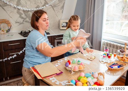 Wide view of little girl plays with a rabbit glove while sitting at the kitchen table with her mom. Child is playing Easter games. Easter Spring Festival 136539920