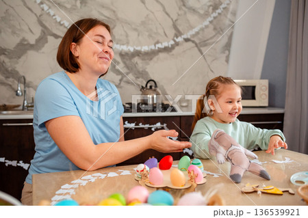 Smiling mother and happy little girl play together with rabbit glove, sitting at Easter table. Spring holiday 136539921