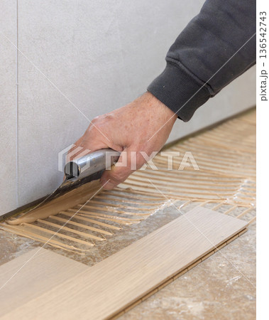 Worker applying parquet flooring adhesive with notched trowel during interior renovation 136542743