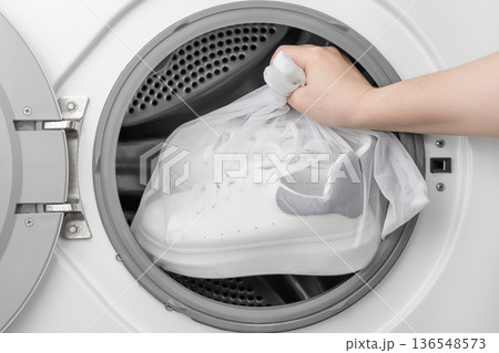 A woman's hand places white sports sneakers into a special laundry bag in the washing machine 136548573