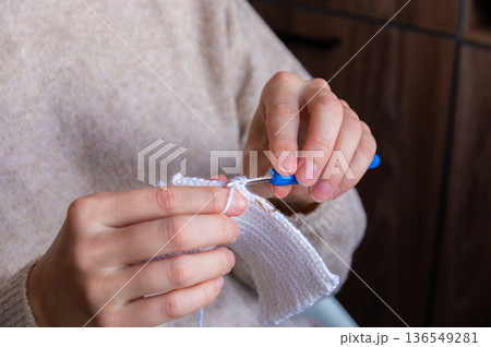Female artisan with light skin using a blue crochet hook to create a white fabric piece while seated indoors with wooden background elements visible Female artisan with light skin using a blue crochet hook to create a white fabric piece while seated indoors with wooden background elements visible 136549281