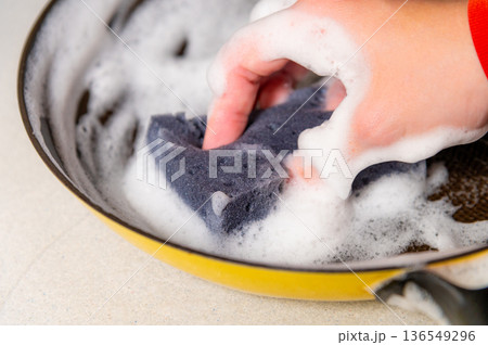 Hand scrubbing a soapy sponge in a yellow frying pan filled with bubbles on a light countertop, showcasing kitchen cleaning activity and dishwashing essentials Hand scrubbing a soapy sponge in a yellow frying pan filled with bubbles on a light countertop, showcasing kitchen cleaning activity and dishwashing essentials 136549296