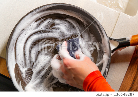 Female Hand of a person wearing an orange long-sleeve shirt scrubbing a black frying pan filled with soapy water on a kitchen countertop with a sink in the background Female Hand of a person wearing an orange long-sleeve shirt scrubbing a black frying pan filled with soapy water on a kitchen countertop with a sink in the background 136549297