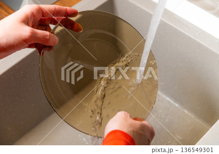 Person washing a transparent glass bowl under running water in a modern kitchen sink with a gray countertop and wooden accents visible in the background Person washing a transparent glass bowl under running water in a modern kitchen sink with a gray countertop and wooden accents visible in the background 136549301