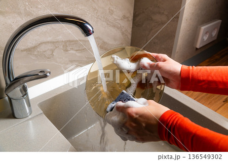 Person with orange long-sleeve shirt washes a round golden plate under running water in a modern kitchen sink with a sleek faucet and textured wall Person with orange long-sleeve shirt washes a round golden plate under running water in a modern kitchen sink with a sleek faucet and textured wall 136549302