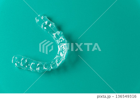 Clear dental aligner placed on a solid turquoise background, showcasing its transparent design and unique shape for orthodontic treatment and teeth straightening 136549316