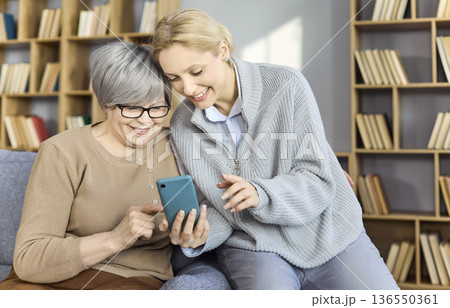 Happy daughter shows grandmother phone features during warm home video call time Happy daughter shows grandmother phone features during warm home video call time 136550361