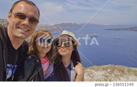 Sunny family selfie with panoramic view of Santorini caldera and Aegean Sea cliffs in bright Mediterranean daylight 136551149