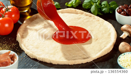Closeup of spreading tomato sauce on fresh pizza dough. Spoon moves gently across the surface, highlighting texture, freshness and authentic Italian pizza preparation. 136554360