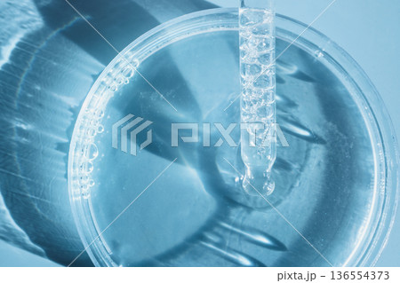 Pipette dropping clear liquid into petri dish with bubbles on blue background 136554373