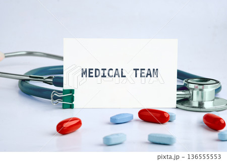 White card reading MEDICAL TEAM stands next to a blue stethoscope. Scattered red and blue pills lie on a bright white surface, symbolizing medical services White card reading MEDICAL TEAM stands next to a blue stethoscope. Scattered red and blue pills lie on a bright white surface, symbolizing medical services 136555553