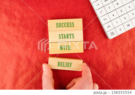 A hand places the final wooden block with BELIEF to form the phrase SUCCESS STARTS WITH BELIEF. It sits on a red textured surface, with a calculator nearby A hand places the final wooden block with BELIEF to form the phrase SUCCESS STARTS WITH BELIEF. It sits on a red textured surface, with a calculator nearby 136555554