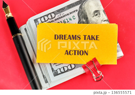 Motivational note Dreams Take Action rests on hundred dollar bills and a writing pen, secured by a red clip on a vibrant red background 136555571