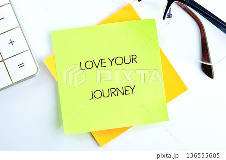 A sticky note with the text love your journey sits on a desk next to a calculator and a pair of glasses, showing an organized workspace 136555605