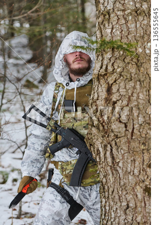 Armed Soldier In Winter Camouflage Peeking From Behind Tree With Rifle And Knife In Snowy Forest 136555645