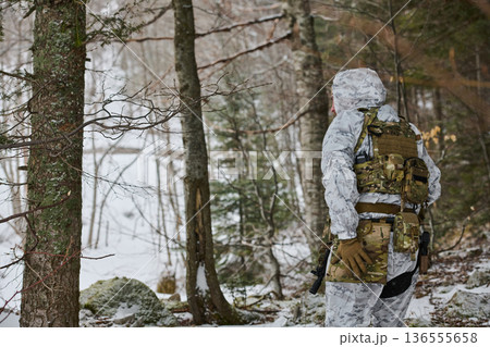 Soldier In White Winter Camouflage Patrols Snowy Forest With Tactical Gear, Backpack And Rifle 136555658