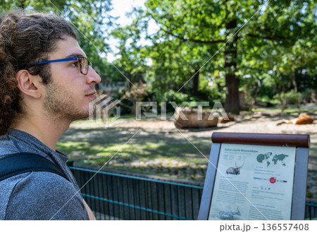 Berlin, germany, august 14, 2023. Young man with glasses reads an information board at berlin zoo, learning about native habitats, wildlife and conservation outdoors 136557408