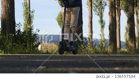 Person rides scooter along a tree-lined path beside a body of water during morning light in a calm outdoor setting 136557584