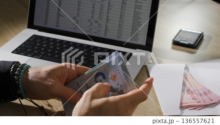 Person counts cash next to a laptop and smartphone on a wooden table during the day in a home office setting 136557621