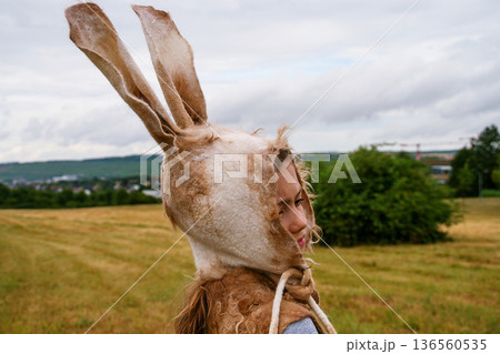 Outdoor side view of a young girl wearing an easter rabbit hat with long ears, standing in a grassy field 136560535