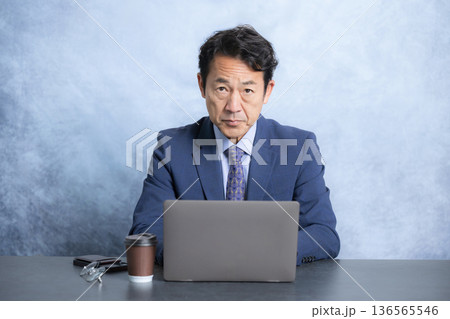 デスクで座りパソコンの前からカメラ目線でスーツで真剣な表情で睨む社長や経営者のビジネスマン 136565546
