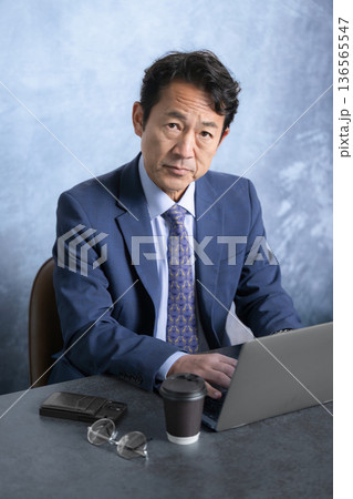 デスクで座りパソコンの前からカメラ目線でスーツで真剣な表情で睨む社長や経営者のビジネスマン 136565547